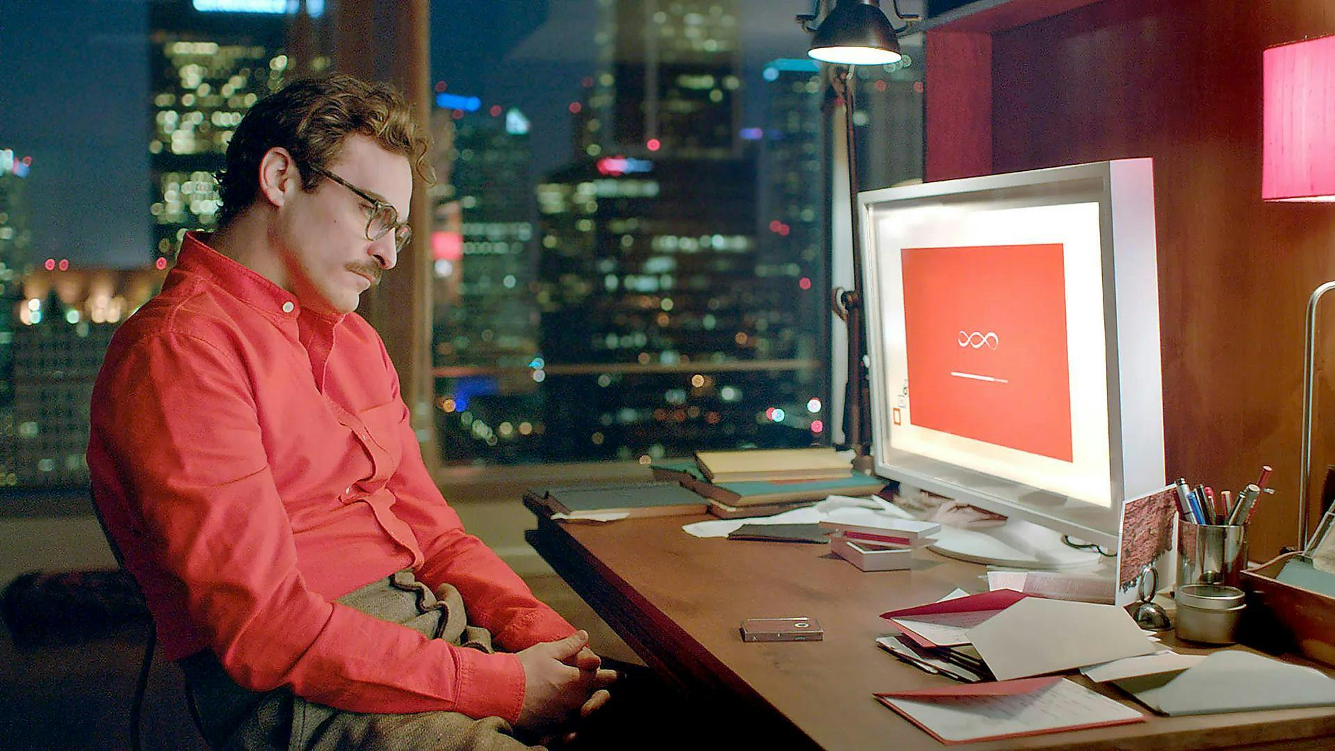 en mann i en rød skjorte sitter ved et skrivebord foran en datamaskin. Bilde fra filmen "Her"